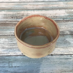 Puede incluir: Un pequeño cuenco de cerámica marrón claro con un borde ligeramente irregular. El cuenco tiene un interior liso y brillante y un exterior rugoso y texturizado.