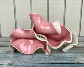 May include: A ceramic sculpture of pink and white abstract shapes, resembling petals or waves. The sculpture has a glossy finish and is arranged in a layered, organic form.