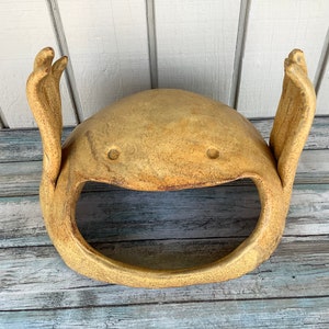 May include: A ceramic planter shaped like a smiling face with two raised arms. The planter is a light brown colour with a speckled texture.