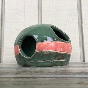 Puede incluir: Una esfera de cerámica verde con tres agujeros y una franja rosa. La esfera está esmaltada y tiene una superficie texturizada.