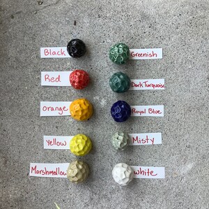 May include: A collection of 10 small ceramic balls in various colors, including black, red, orange, yellow, marshmallow, white, greenish, dark turquoise, royal blue, and misty. Each ball has a textured surface.