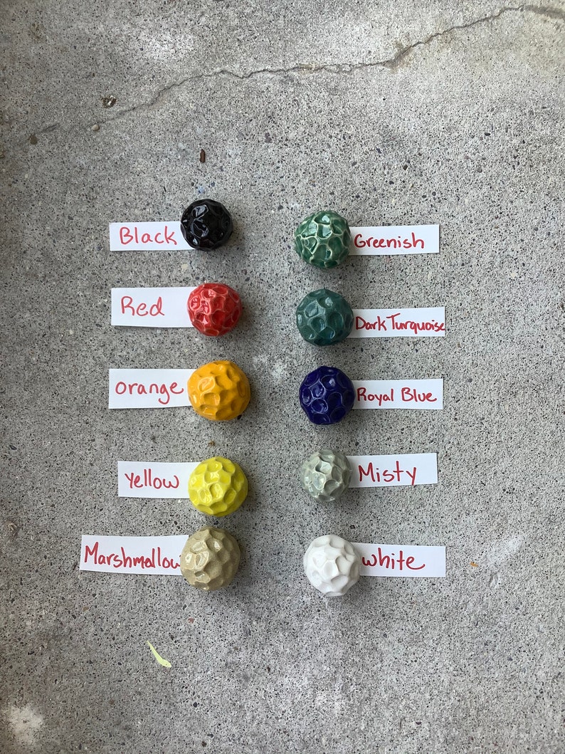 May include: A collection of 10 small ceramic balls in various colors, including black, red, orange, yellow, marshmallow, white, greenish, dark turquoise, royal blue, and misty. Each ball has a textured surface.
