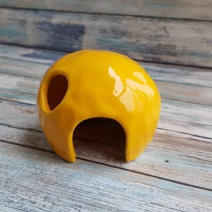 May include: A yellow ceramic hide with a rounded shape and a large opening. The hide has a smooth, glossy finish.