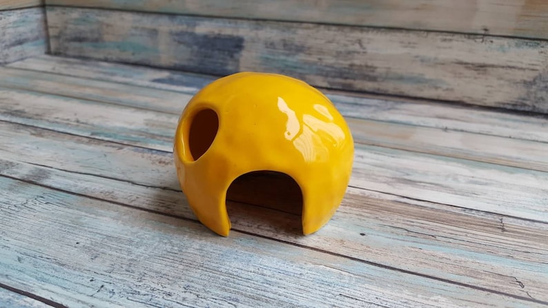 May include: A yellow ceramic hide with a rounded shape and a large opening. The hide has a smooth, glossy finish.