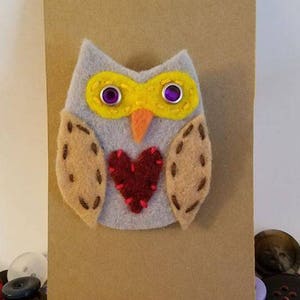 Könnte beinhalten: Eine Filz-Eulenbrosche mit gelbem Schnabel, gelben Augenbrauen, lila Augen und einem roten Herzen auf der Brust. Die Eule ist an einem braunen Papieretikett mit einem weißen Kreis befestigt.