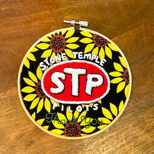 May include: Embroidered hoop art featuring a design with a red and white STP logo, surrounded by yellow sunflowers on a black background. The text "STONE TEMPLE PILOTS" and "Thank You" are also included.