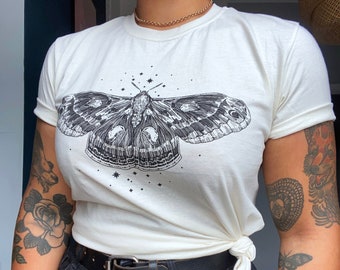 May include: Cream-colored t-shirt featuring a detailed black and white moth illustration with surrounding stars. The shirt is knotted at the waist and paired with black jeans and a black belt. A gold chain necklace is visible.