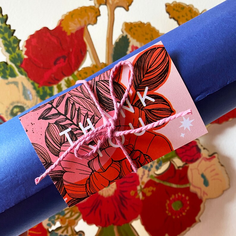 May include: A pink and red thank you card with a floral design and a pink string tied around it. The card is on top of a blue rolled up poster.