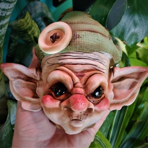 May include: A ceramic gnome head with a green hat and a brown button. The gnome has a grumpy expression with large eyes and a prominent nose.