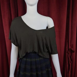 May include: A dark grey, off-the-shoulder top with a V-neck and short sleeves. The top has a slightly distressed, raw hemline and a small pocket on the front. It is paired with a pleated plaid skirt in navy, green, and yellow.
