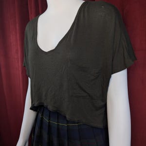 May include: Dark olive green crop top with a deep V-neck and a small pocket on the chest. The top has short sleeves and a raw hem. The top is paired with a plaid skirt.