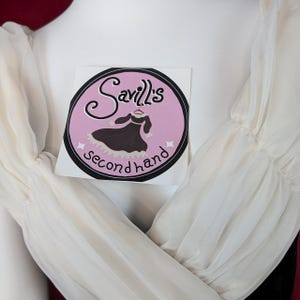 May include: Close-up of a white dress with sheer sleeves. A sticker with the text "Savill's Secondhand" is on the dress. The sticker is round with a pink background and a black border. The dress has a black bodice.