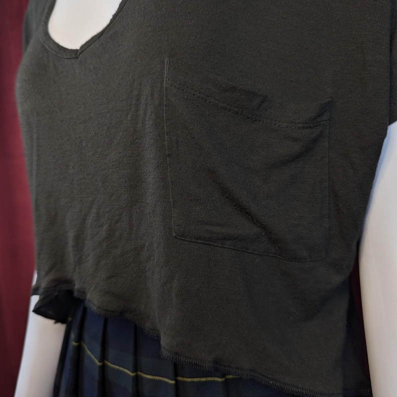 May include: Dark green, cropped t-shirt with a V-neck and a pocket on the chest. The shirt has a raw hem and is paired with a plaid skirt. The fabric appears to be a soft, lightweight material.