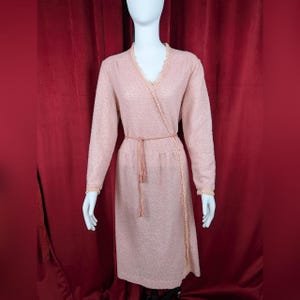 May include: A long-sleeved, knee-length wrap dress in a soft, textured pale pink fabric. The garment features a V-neckline trimmed with delicate lace, long sleeves with lace-trimmed cuffs, and a matching thin tie belt at the waist.