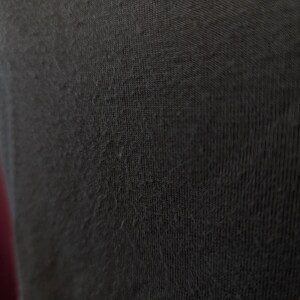 May include: Close-up of a dark grey fabric with a textured surface. The fabric appears to be a knit or woven material, with small fibres visible on the surface. The image highlights the texture and colour of the textile.