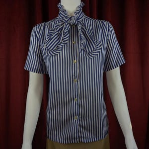 May include: A short-sleeved blouse with vertical blue and white stripes. The blouse features a ruffled collar and a bow tie detail at the neckline. Gold-colored buttons run down the front.