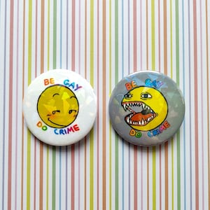 May include: Two round buttons with a white background and rainbow text. The text on the buttons reads "Be Gay Do Crime". One button features a yellow smiley face with a mischievous expression. The other button features a yellow cartoon monster with a large open mouth and sharp teeth.