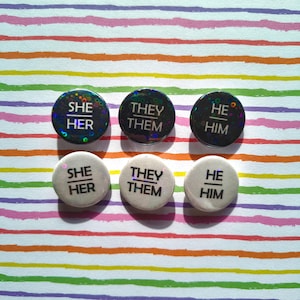 May include: Six circular pronoun badges, each displaying "SHE HER", "THEY THEM", or "HE HIM". Three have a holographic effect, and three are white. They are set against a backdrop of colourful horizontal stripes.