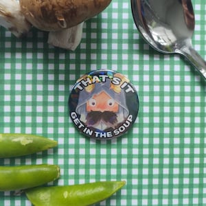 Op de afbeelding: Een ronde knop met een cartoonfiguur dat een capuchon draagt en de tekst "That's it, get in the soup".