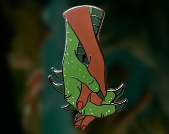 May include: A green and brown enamel pin depicting a human hand holding a green, scaly hand. The green hand has white spots and red fingernails.