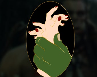 May include: A gold-colored pin with a black background depicting two hands intertwined. One hand is green and the other is a pale skin tone with red nail polish.
