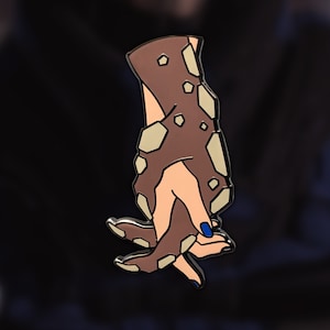 May include: A brown and beige enamel pin of two hands holding each other. The hands have a textured, rocky surface. The nails are painted blue.