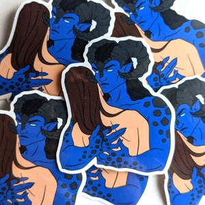 May include: A set of stickers featuring a blue-skinned, horned creature with black horns and a black and blue patterned body. The creature is embracing a person with long brown hair. The stickers are printed on white paper.