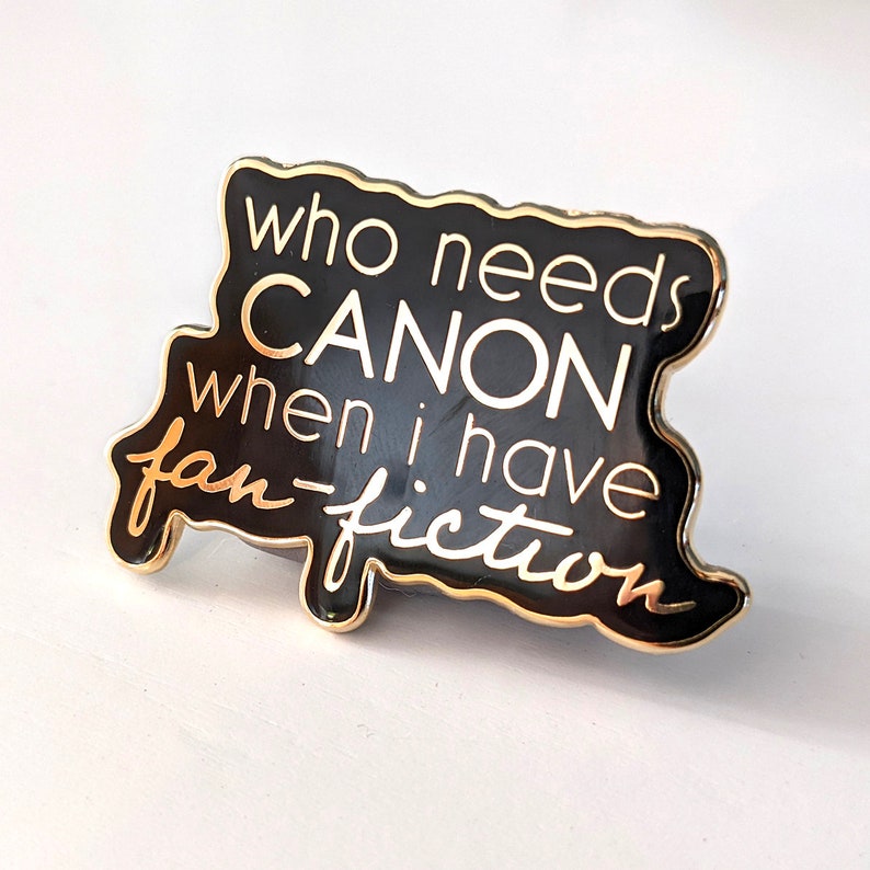 Puede incluir: Pin de esmalte negro y dorado con el texto "&iquest;Qui&eacute;n necesita canon cuando tengo fan-fiction?"