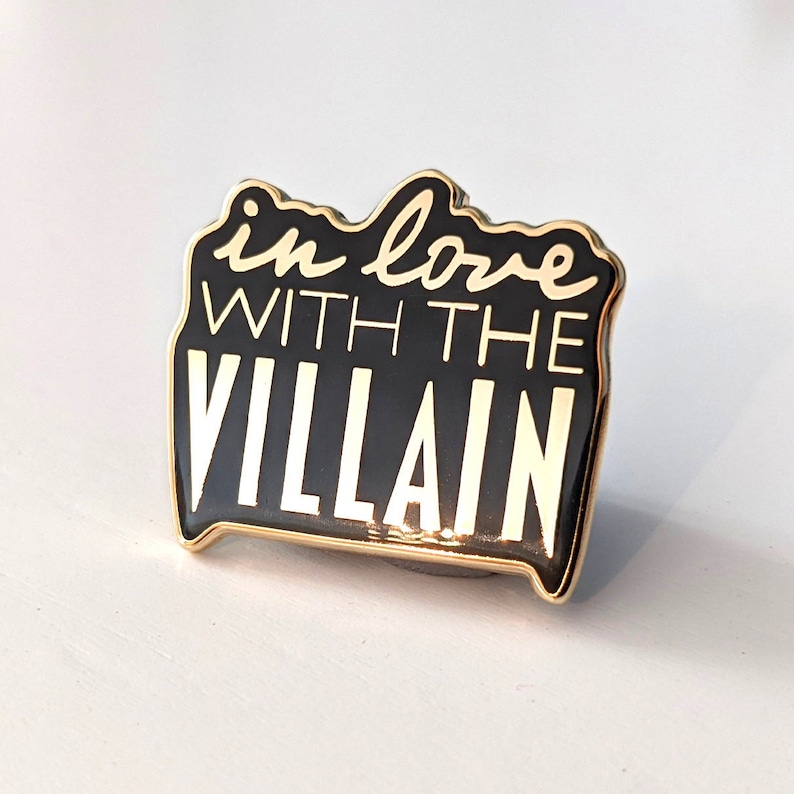 May include: A black enamel pin with gold trim, featuring the phrase "in love with the villain" in white script and block letters. The pin has a rectangular shape with rounded edges.