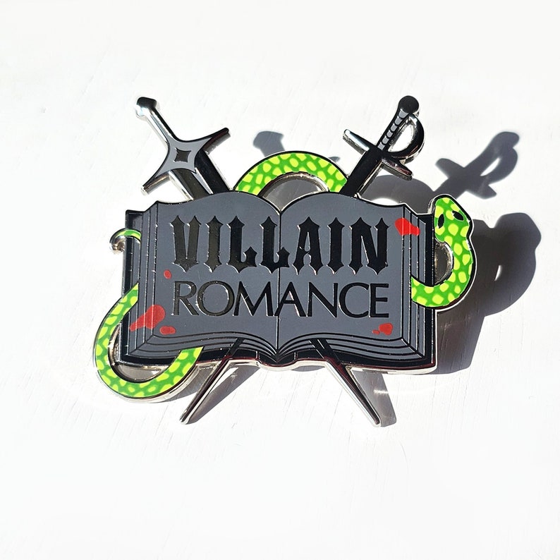 May include: A silver metal pin with a green snake wrapped around a closed book. The book has the words "Villain Romance" printed on the cover.