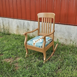 May include: A wooden rocking chair with a blue and brown patterned cushion. The chair is sitting on a patch of green grass in front of a red metal wall.