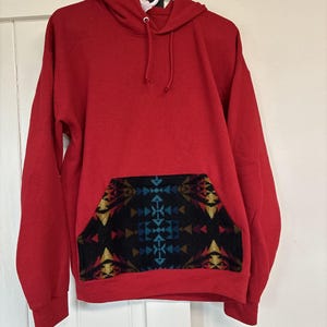 May include: A red hooded sweatshirt with a patterned pocket. The pocket features a geometric design in shades of black, blue, yellow, and gold. The sweatshirt is hanging on a white hanger.