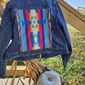 May include: A denim jacket with a colorful patterned back panel. The panel features vertical stripes in shades of pink, blue, yellow, and white with geometric designs. The jacket is displayed outdoors with a white pumpkin and dried corn stalks.
