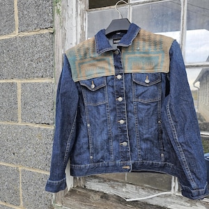 May include: A dark blue denim jacket with a patterned shoulder yoke in shades of tan, teal, and blue. The jacket has a button-up front, two chest pockets, and is hanging on a black hanger.