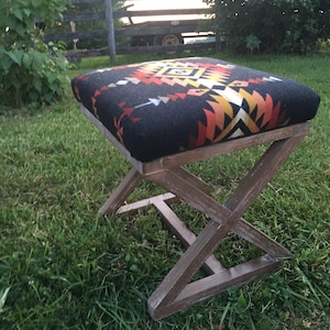 May include: A square ottoman with a dark gray patterned top and a wooden X-frame base. The top features a geometric design in shades of red, orange, yellow, and white. The wooden base has a weathered appearance.