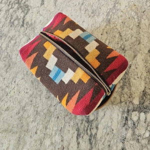 May include: A rectangular, zippered pouch with a colorful geometric pattern in shades of brown, red, orange, yellow, blue, and white. The zipper is gold-toned, and the pouch appears to be made of a woven fabric.
