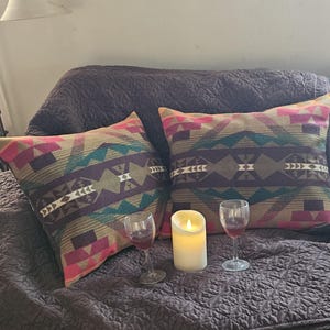 Puede incluir: Dos cojines decorativos con un patrón geométrico en tonos rosa, turquesa, marrón y morado. Una vela encendida y dos copas de vino se colocan sobre una manta texturizada marrón oscuro.