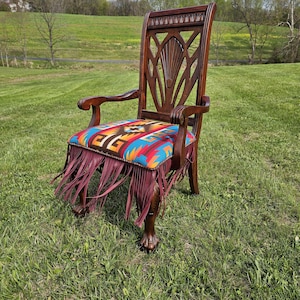 May include: A dark wood armchair with a colorful patterned seat cushion and burgundy fringe. The chair has carved details on the back and arms, and is set on a grassy lawn.