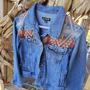 May include: A blue denim jacket with a patterned design on the shoulders. The jacket has a classic collar, button closures, and two chest pockets. The shoulder design features geometric shapes in shades of orange, brown, and blue. The brand name "J.CREW" is visible on the label.