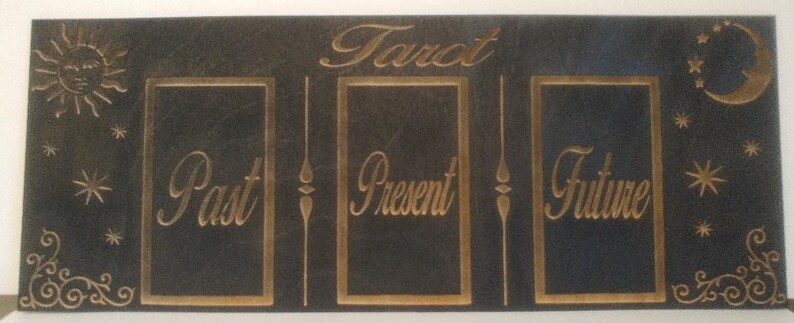Puede incluir: Un tablero de madera negro con letras doradas que dicen "Tarot" y tres ranuras etiquetadas como "Pasado", "Presente" y "Futuro".