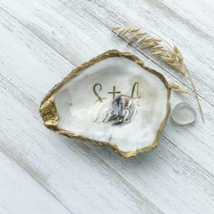 May include: An oyster shell with a gold-painted rim and the initials "S + A" and the date "9.23.22" in gold script. The shell sits on a white wooden surface, with a sprig of wheat and a small, smooth stone nearby.