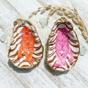 May include: Two decorative oyster shells with gold edges. Each shell features a tiger design. One tiger is orange, the other is pink, both set against a zebra-striped background. A unique home decor item.