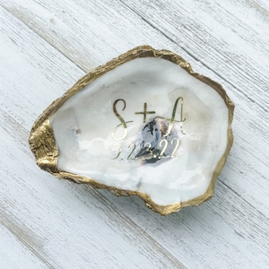 May include: A white oyster shell ring dish with a gold rim. The shell is decorated with the text "S+A 9.23.22" in gold lettering.