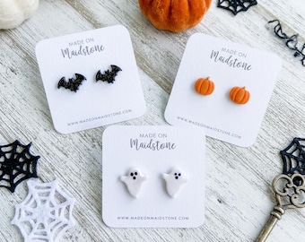 May include: Three pairs of stud earrings on white display cards labeled MADE ON MAIDSTONE. One pair features black glitter bats, one pair features orange pumpkins, and one pair features white ghosts with black eyes and mouths on a light surface.