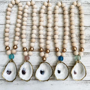 May include: Five decorative beaded garlands with oyster shell pendants. Each garland features natural wood beads, gold-toned accents, and a colorful glass bead. The oyster shells have a gold-colored trim and a dark interior.