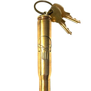 Puede incluir: Un llavero con forma de bala de latón con un diseño de calavera. El llavero contiene dos llaves en un anillo de latón. La carcasa de la bala es de color dorado y tiene un emblema de calavera detallado.