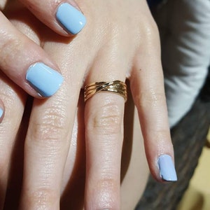 Könnte beinhalten: Ein goldener Ring mit geflochtenem Design. Der Ring wird am Ringfinger einer Hand mit hellblauem Nagellack getragen.