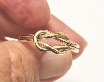 May include: A gold-colored ring in a knot design. The ring is held between fingers, showcasing the intricate knot detail. The ring's band is smooth and simple, complementing the knot's complexity. The ring is likely made of gold or a gold-colored metal.