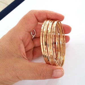 Op de afbeelding: Een set van acht gouden armbanden met een gehamerde textuur. De armbanden zijn op elkaar gestapeld en worden in een hand gehouden.