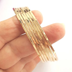 Puede incluir: Conjunto de ocho pulseras de brazalete martilladas de color dorado. Las pulseras están apiladas una encima de la otra y están dispuestas en forma de curva.